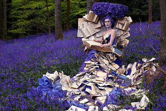 woman in dress covered with pages of books with purple headdress and surrounded by purple flowers