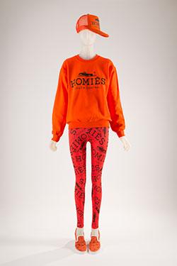 bright orange ensemble; sweatshirt with black printed Homies South Central logo, printed leggings, baseball style cap, and slip-on sneakers