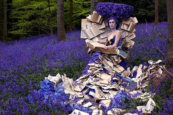woman in a violet-blue dress covered with pages of books with the same color headdress and surrounded by bluebell flowers in the clearing of the woods