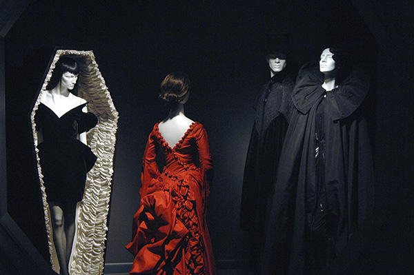 four mannequins on display, from left to right: a mannequin in a velvet black dress in a coffin, a red ruffled dress, and two mannequins in black capes and ensembles