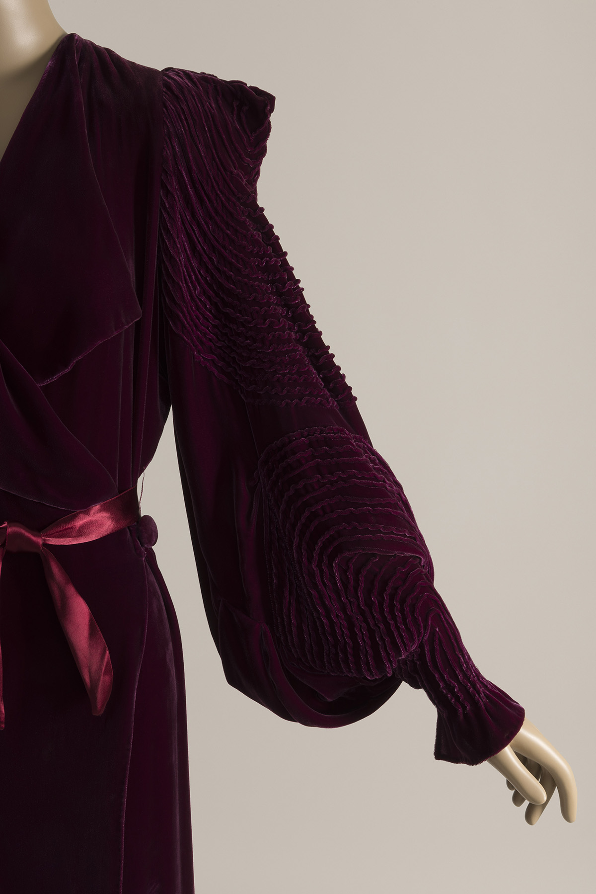Partial side view of a woman’s dressing gown in velvet, tied at the waist with a matching silk ribbon. The full sleeve has a sharply defined shoulder, a tapered cuff, and elaborate pin-tucks in concentric circles.