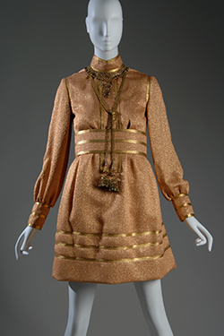 short cocktail dress with gold braid collar, placket, cuffs and hem, and attached brass filigree necklace with pendant tassels and wide belt