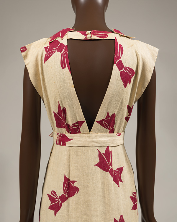 Back view of an ivory linen dress with printed red bows. The open back comprises of an upside down triangle, with the tip ending at the waist.