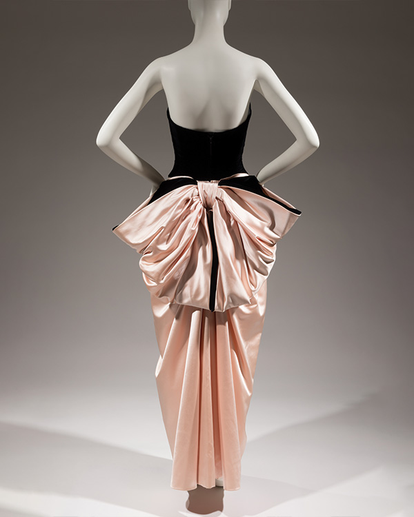 Back view of a black velvet and pink satin dress, with a large pink satin bow tied at the lower waist. 