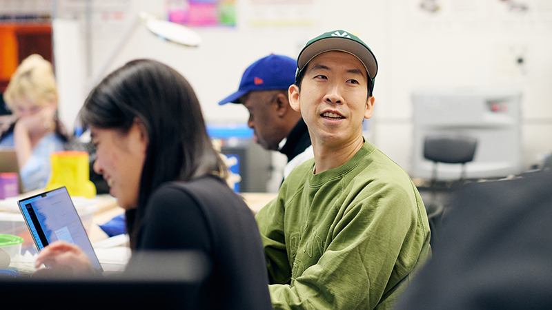 An Asian male student looking off into the distance surrounded by other students working on computers