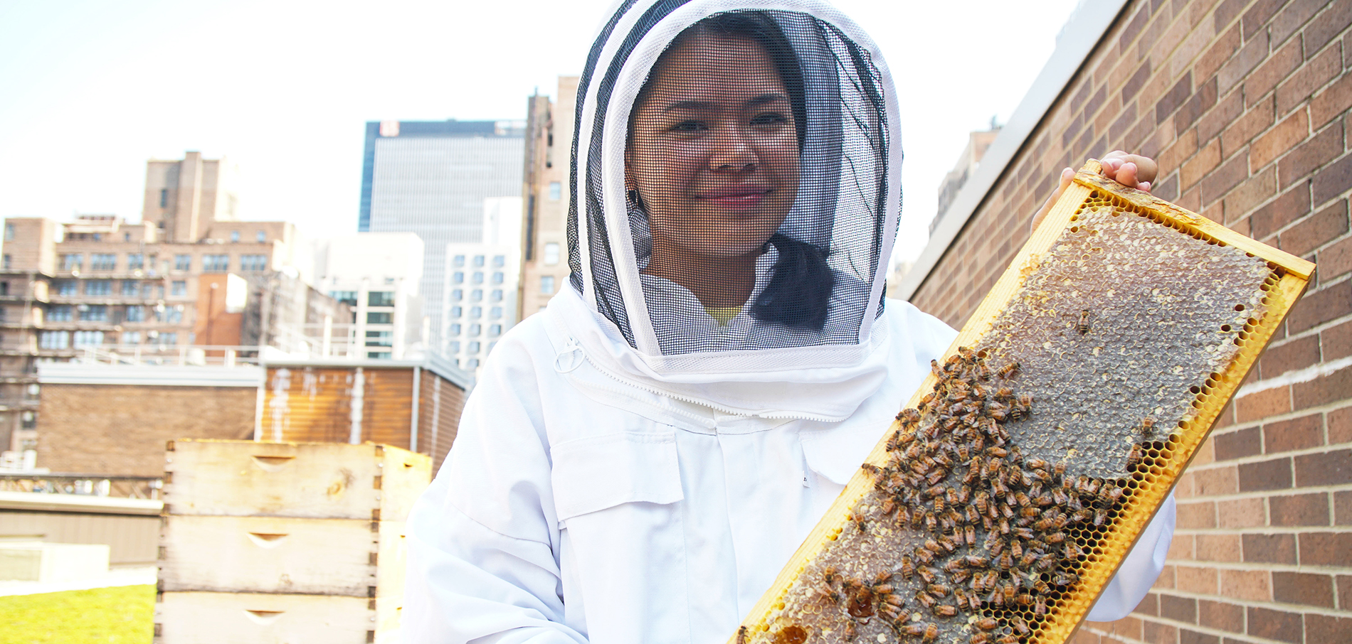 A female student with dark hair in beehive protective gear is holding a comb dripping with honey