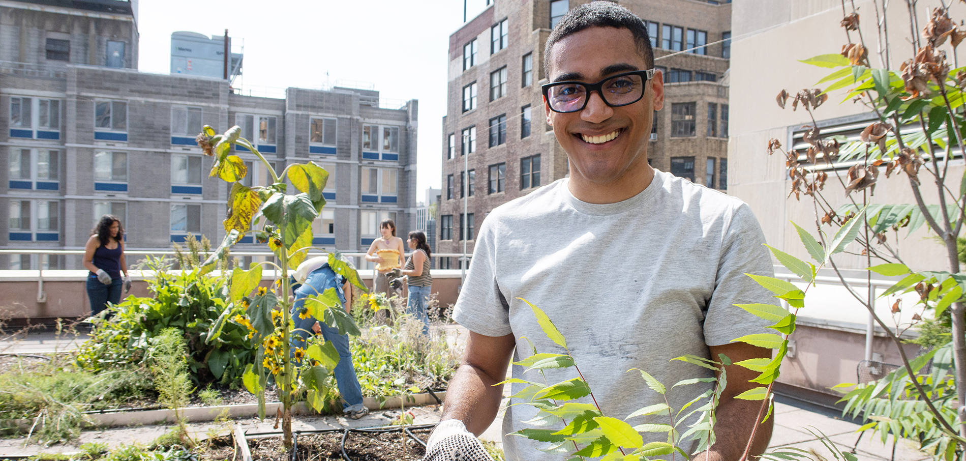 A male student wearing a white t-shirt and black framed glasses is smiling on top of the roof next to the dye garden