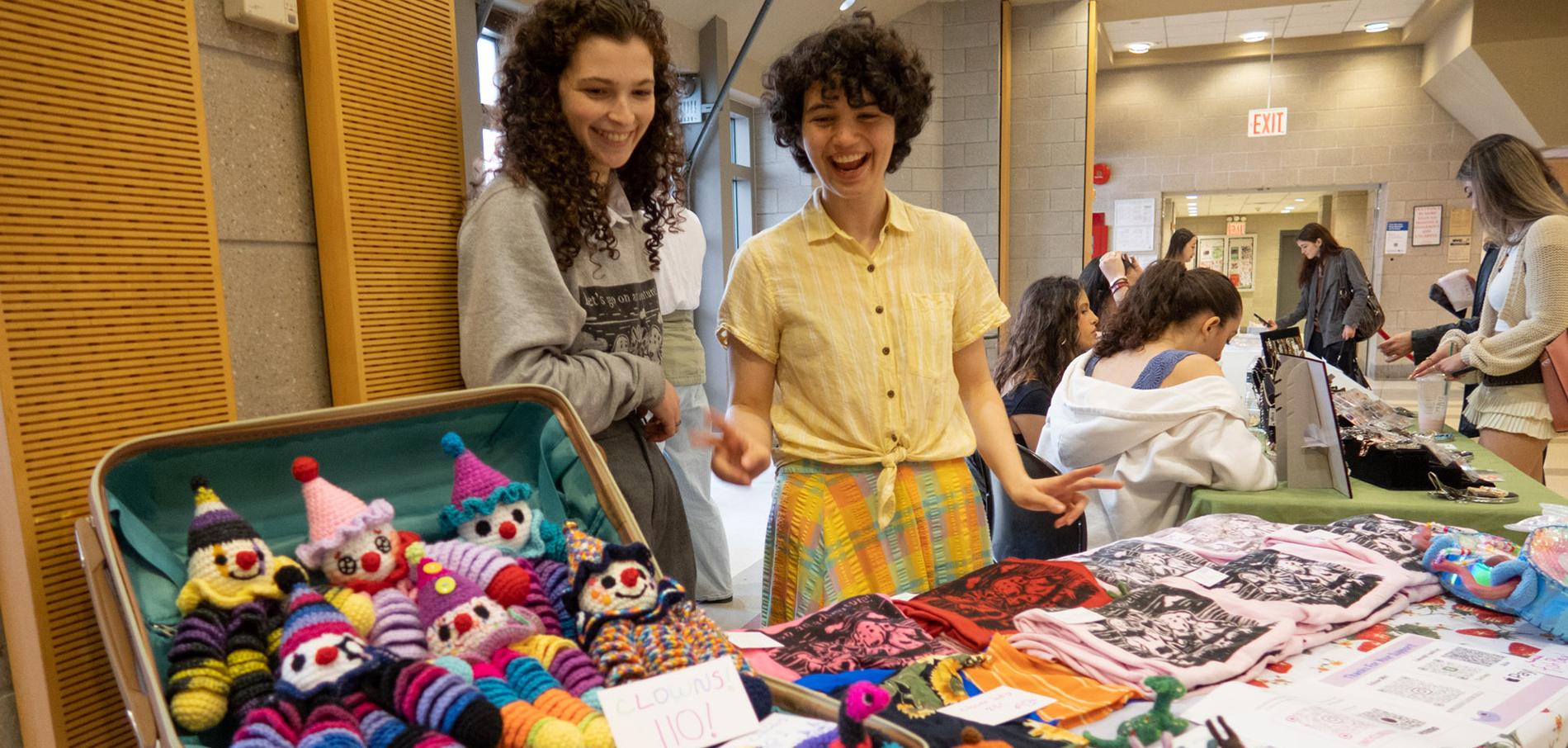 Two female students standing behind a table of crafts they are selling such as yarn puppets and screenprinted tshirts.