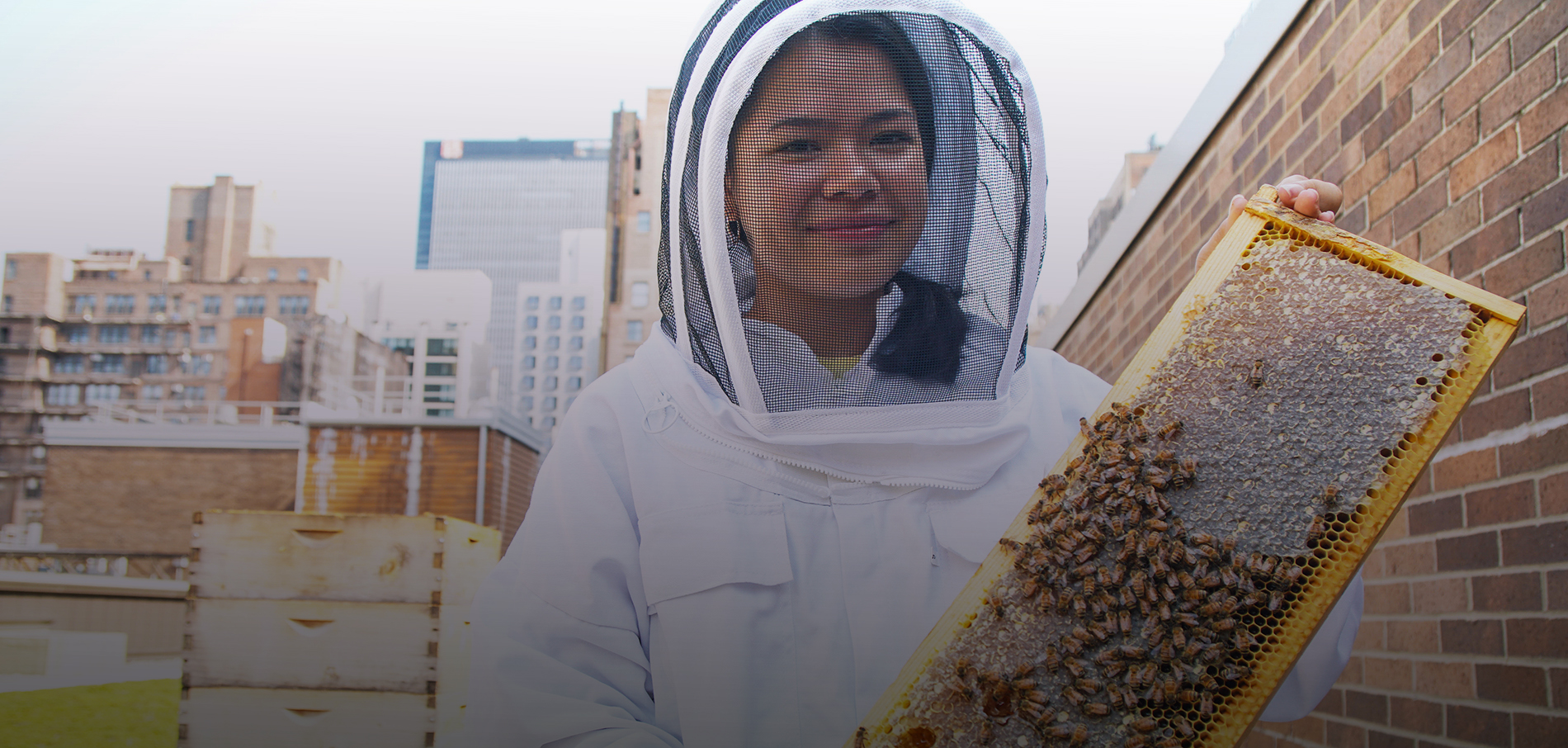 A female student with dark hair in beehive protective gear is holding a comb dripping with honey