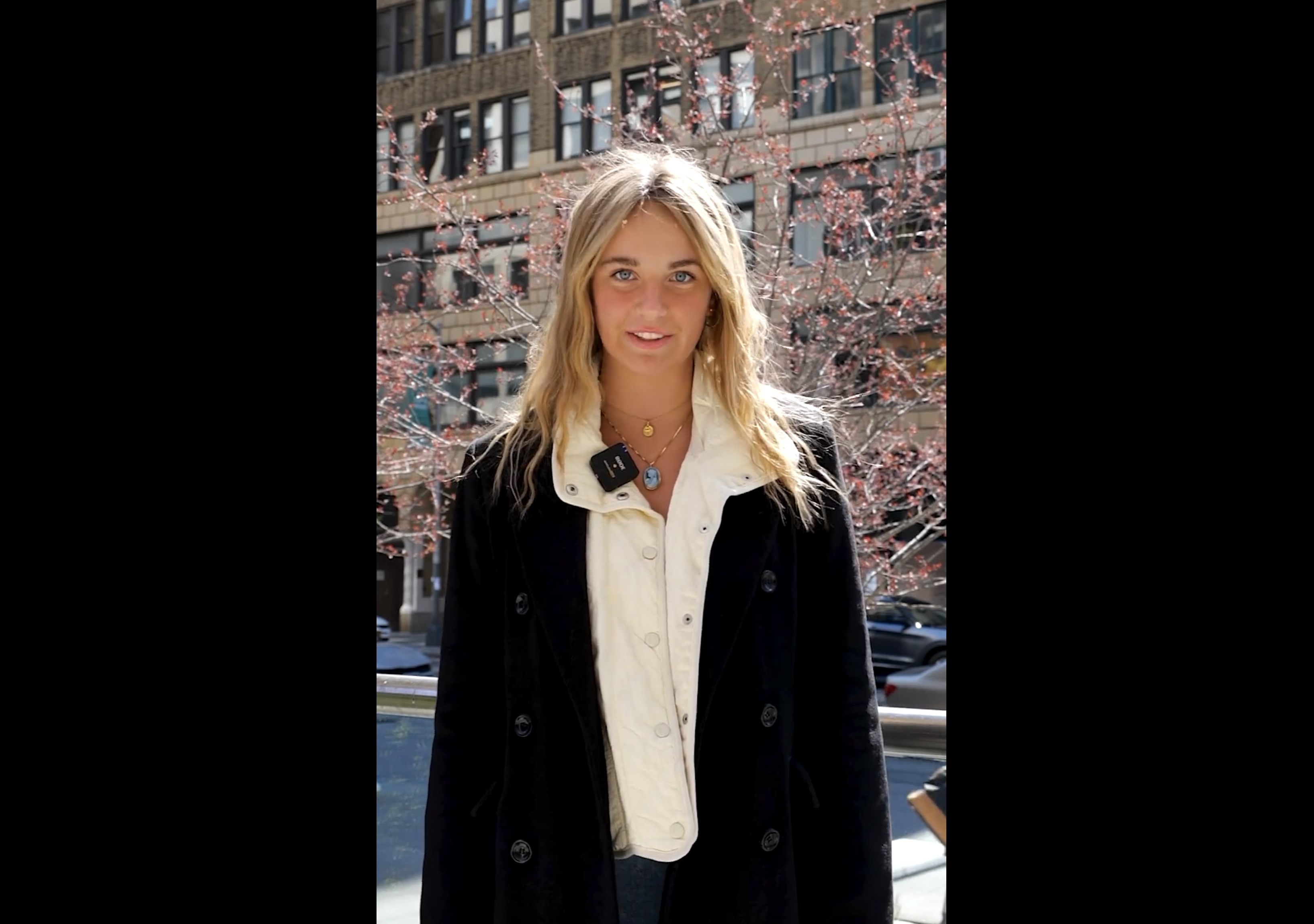 A blonde-haired female student standing outside of FIT with cherry blossom trees behind her.