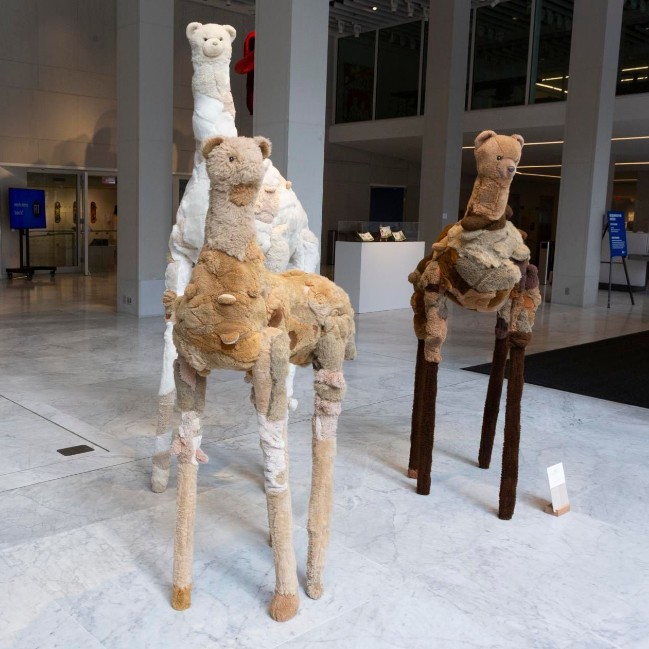 a group of three creatures made of soft, plush patchwork of furlike materials. With the torsos and legs reminiscent of giraffes or llamas with teddy bear heads at the end of elongated necks. One is in sandy and light brown tones, another one in pale creams and whites, and the third in darker browns.