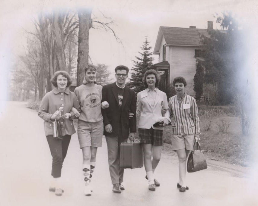 Five smiling people with linked arms walking down a country or suburban road. One man is in the middle, and the women flanking him on both sides in twos. It seems to be either early spring or fall with the vegetation at rest, there are trees lining the road, and a part of the house is visible just behind them. Three women are wearing thigh-length shorts. One woman is wearing a sweatshirt, and the rest are attired in coats and jackets. Women are carrying handbags, and a man is carrying a suitcase.