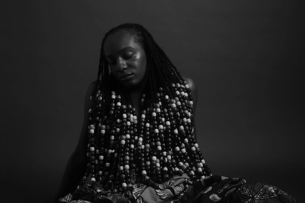 At the center of a black and white horizontally oriented photograph is a figure seated on the floor or the ground. The long hair is styled in dreadlocks that are adorned in large beads shaded from light to dark. The braids are draped to completely cover the upper body and torso with only naked arms that are placed behind for support slightly exposed. A little bit of a Ntoma patterned textile covering the lower body is visible at the bottom completes the image.