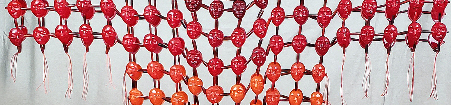 A closeup detail of the war shirt. Neckline and sleeves stretched out. Maroon and red colored large beads shaped like faces sparsely strung in a lattice pattern.