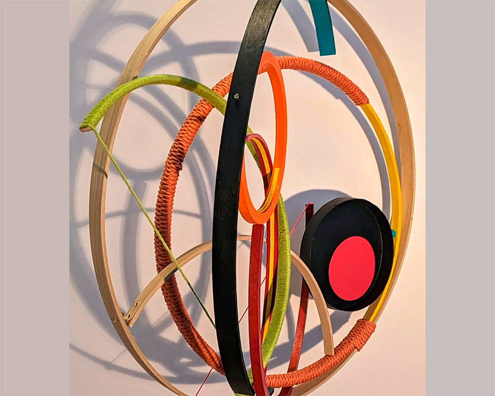 a composition of wood strips bent to form circles and incomplete circles in carious sizes, some painted, some covered with yarn or rope, arranged and held together with metal fasteners or string the piece is hung against the wall, and casting a shadow on the wall creating an intricate pattern of intersecting 3d and 2d lines, shapes, and colors