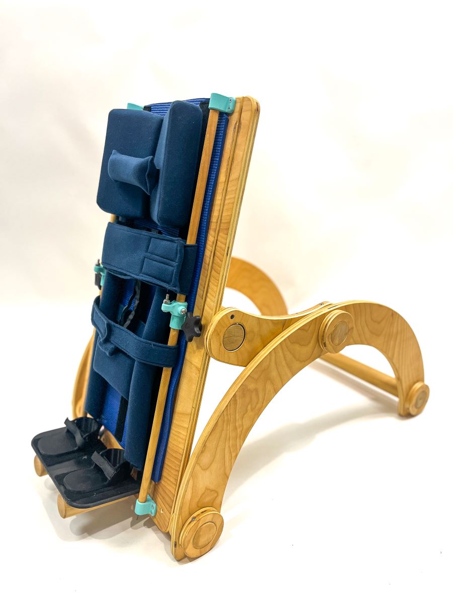 Wood and cardboard children’s standing support device, featuring adjustable back and curved legs, a padded blue seat and head pillows with footrests and adjustable velcro harness.