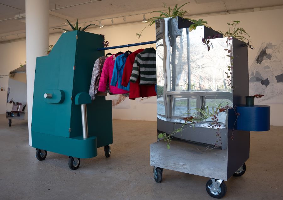 Two mobile closet structures on caster wheels, one teal and one with a mirrored surface, connected by a rack of colorful clothing. Created for mutual aid and clothing distribution to unhoused people. Both units incorporate potted plants.