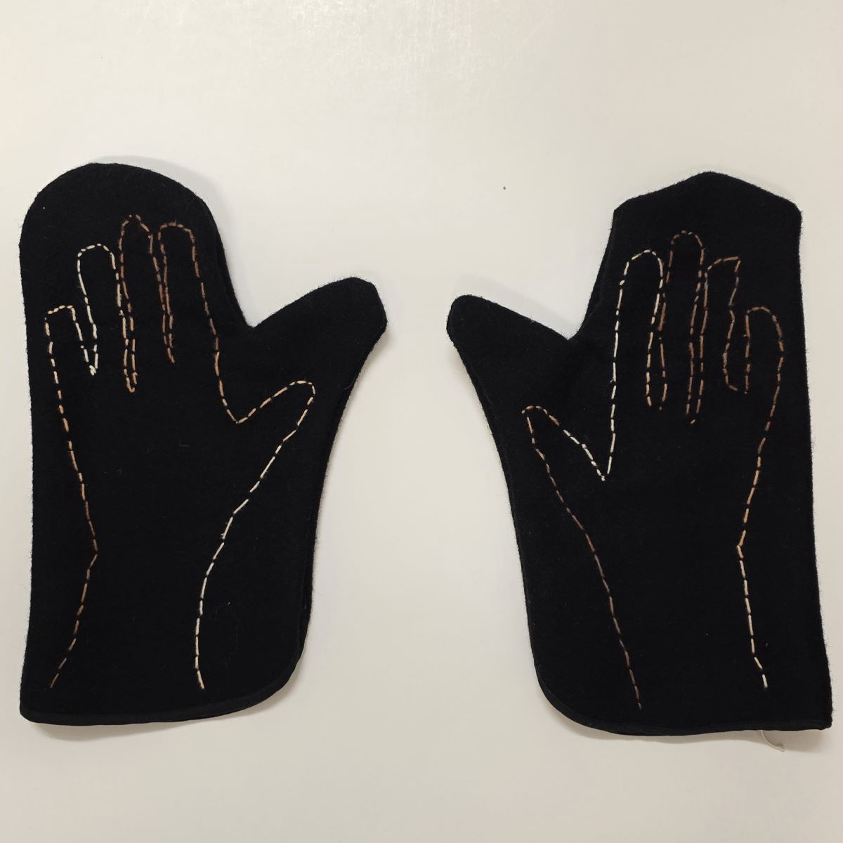A pair of black mittens laid flat on a white surface, each featuring an embroidered outline of a hand in brown and beige stitching on the top side, used in class demonstrations to simulate limited fine motor control—encouraging empathy and reflection on accessibility through the experience of reduced hand dexterity.