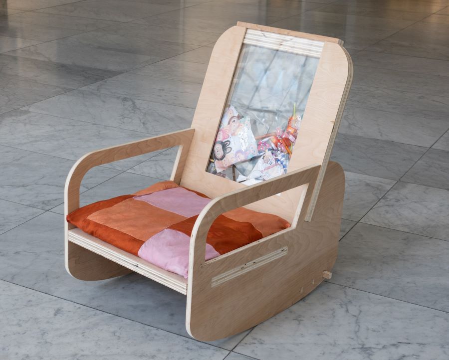 A rocking chair made of light plywood with a patchwork orange and pink cushion. The backrest has a transparent panel filled with plastic packaging materials, highlighting consumption and recycling.