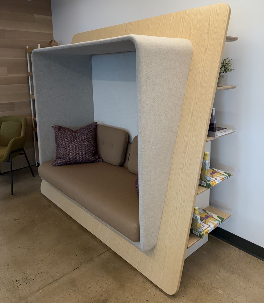 LeanTo is composed of natural wood surfaces with rounded corners, earth-tone upholstery, and built-in shelves along the outer wall. Shelves differ in length because they are affixed to a wooden panel that slants diagonally, from bottom to top corner, framing the structure. Overall, the structure feels soft, calming, and inviting.