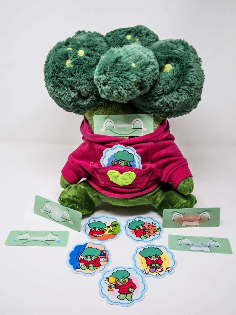Plush toy shaped like a broccoli wearing a pink hoodie, displayed with interchangeable illustrated cards showing different activities and facial emotions. The toy is designed as an interactive aid for children to explore feelings and communication.