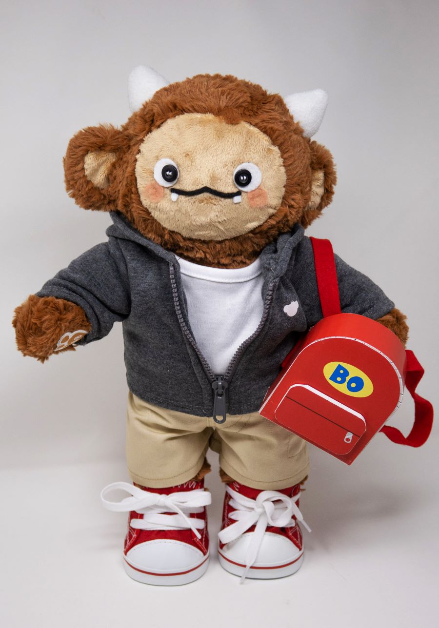 Plush monster doll with brown fur, small horns, and a friendly face, dressed in a gray hoodie, khaki pants, and red sneakers. The toy carries a red backpack labeled 'Bo,' suggesting it is designed as a character-based childhood learning companion.