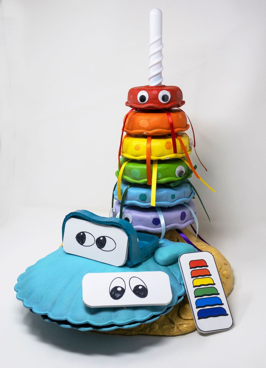 Colorful educational toy for interactive and sensory play, featuring stackable rainbow-colored discs with cartoon eyes and laces, topped with a white spiral handle. A clam-shaped container is in the foreground. It has cartoon eyes and a rectangular piece with bright color swatches.
