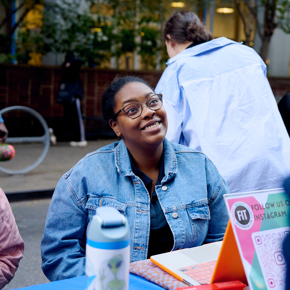 Woman wearing a denim jacket and glasses is tabling on West 27th street, engagedly listening to visitor