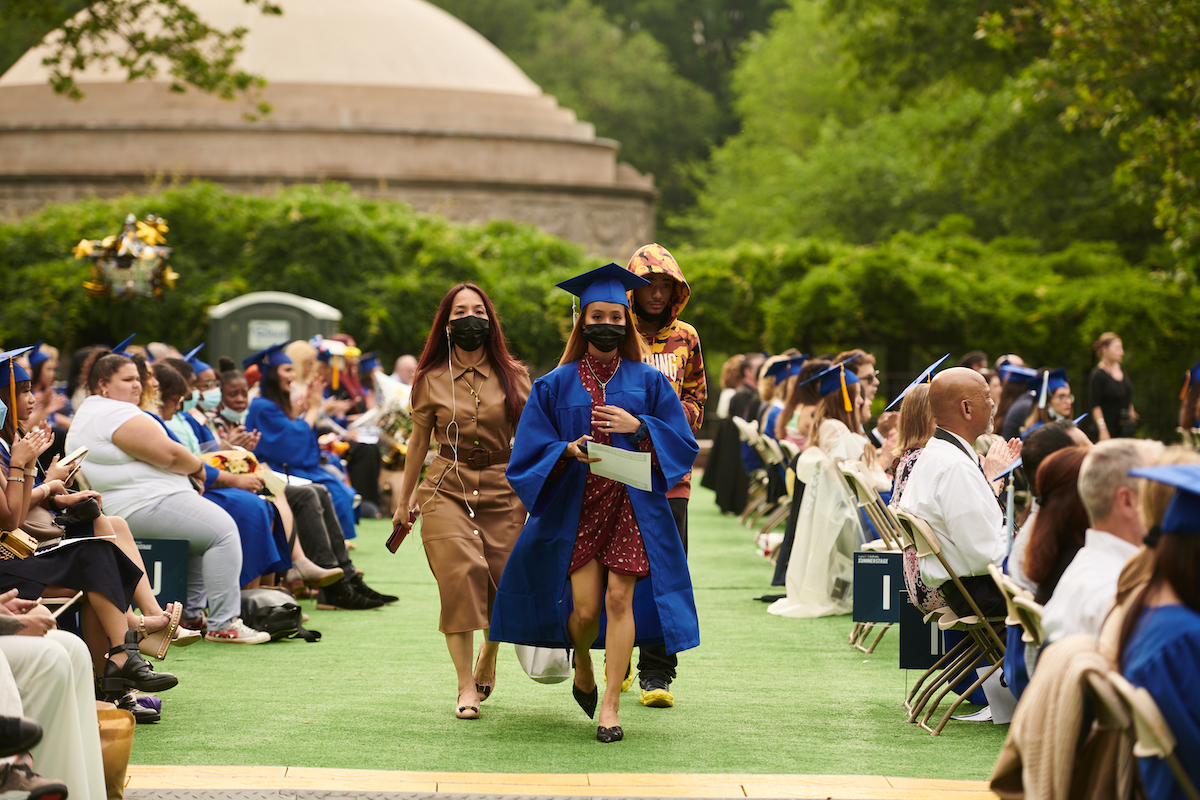 three people, one in cap and gown, walking among seated viewers of commencement