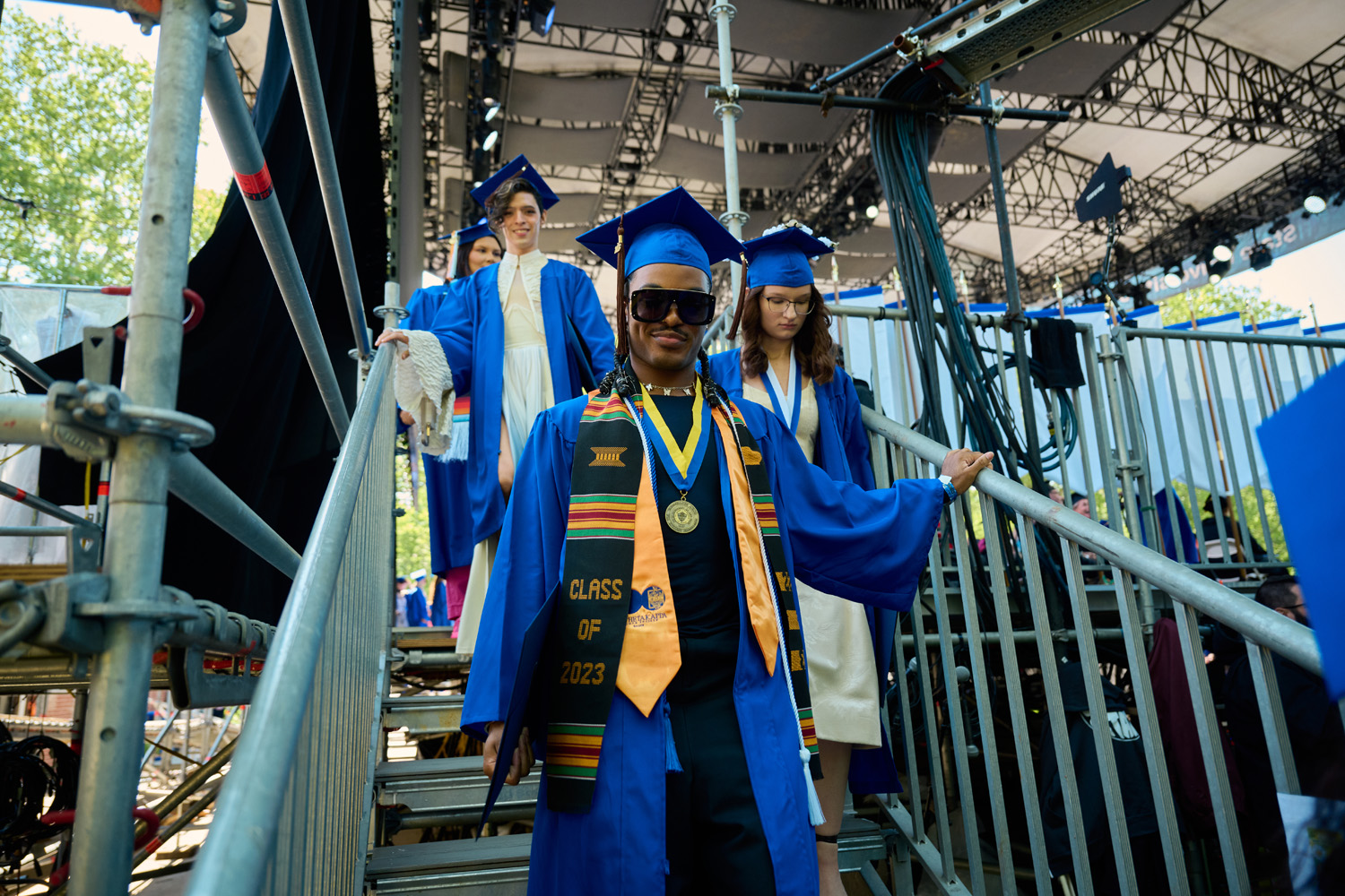 three students in cap and gown walking off commencement stage
