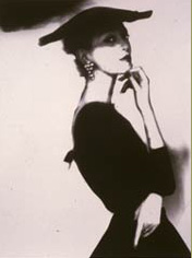 woman in black dress with a black hat turned toards camera with one hand on chin