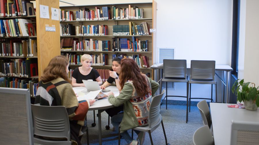 students in the library sitting round a table, surounded by the bookshelves 