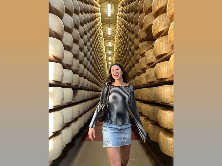 a smiling woman walking through a passageway between a floor to ceiling shelves that are filled with the rounds of cheese as far as the eye can see...