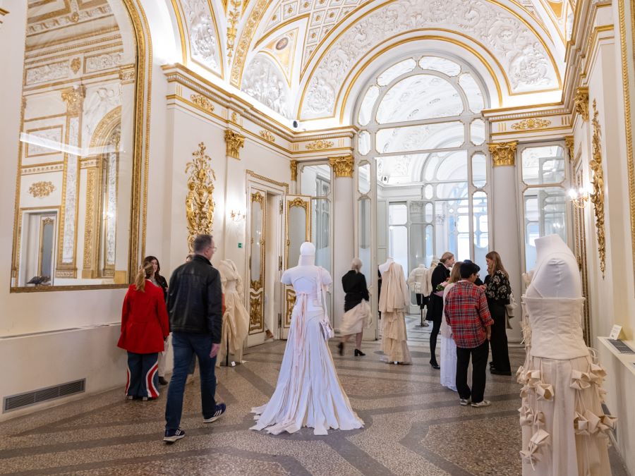 Mannequins dressed in muslins displayed in a gilded, palace-like, light-filled gallery space featuring large mirrors, and high arched doorways. A handful of people are viewing the exhibition.