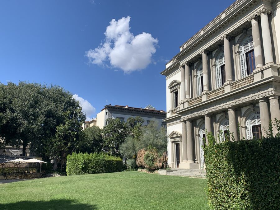 a blue summer sky with a few puffy clouds in view, a large classical building with arched windows and columns is on the right side of the image, a lawn extends in front of it with other greenery, trees and more buildings visible in the background