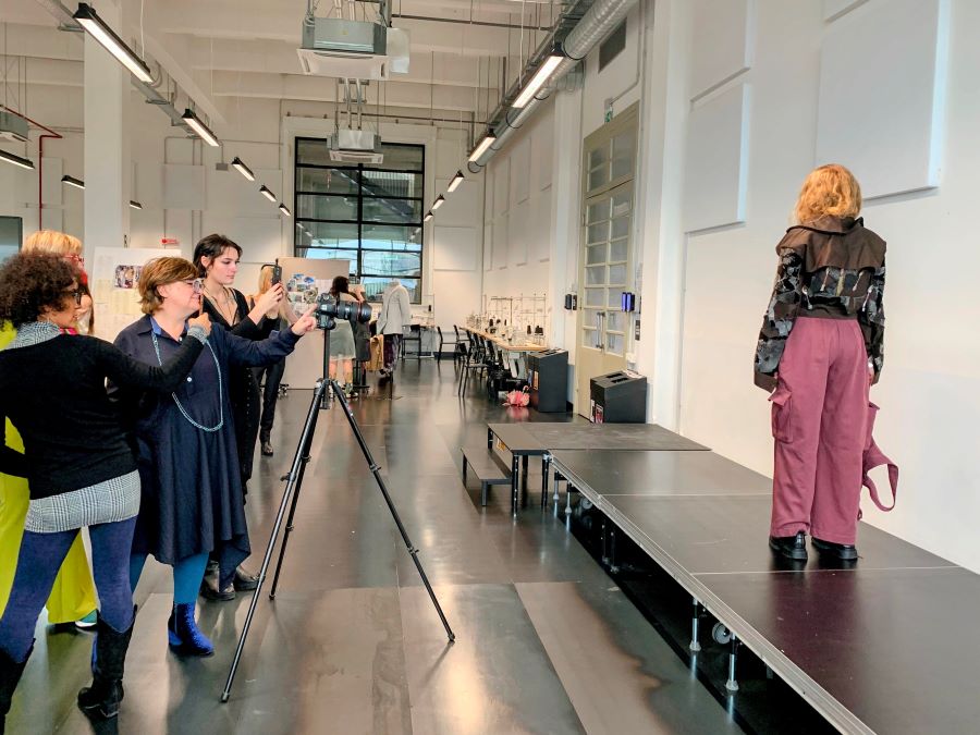 a photo session of a student standing on an elevated platform/runway modeling to a group of women taking pictures. Student's back is turned to the camera. Her outfit is comprised of mauve-pink wide-leg cargo pants, and a patchwork-resembling earth-toned top, with a wide yoke collar. 