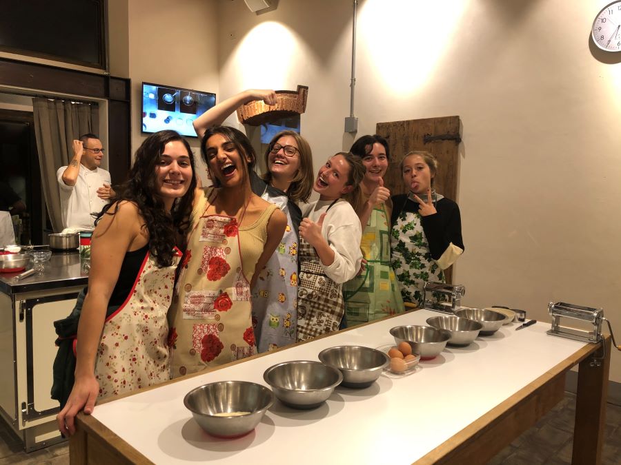 a group of smiling students lined up behind a heavy kitchen work-table. Six empty metal mixing bowls are neatly lined up in the middle of the table; two pasta rollers are attached to the far end of the table. 