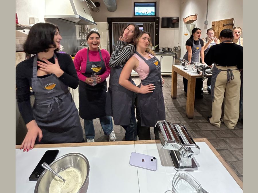 two groups of students wearing aprons are gathered around the two heavy kitchen work tables. A bowl with flour, a pan of water, and a pasta rolling apparatus are on the table closest to the camera. Students seem to be enjoying the activity.