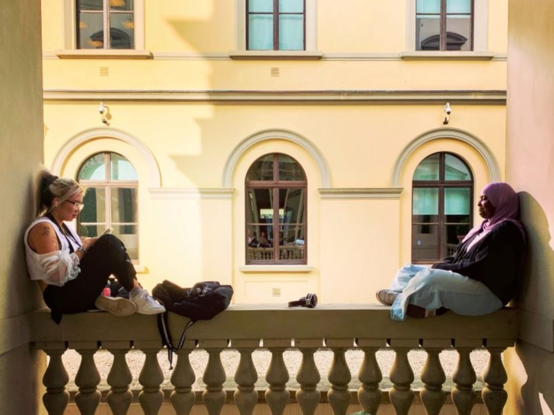 two students sitting on an ornamental parapet, immediately across from a renaissance building with arched windows.