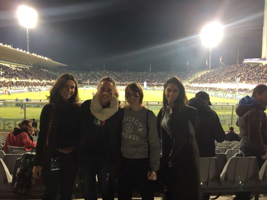 Four students are facing the camera during a sports game. Behind them is a soccer/football stadium full of spectators. Two bright lights are illuminating the evening scene.