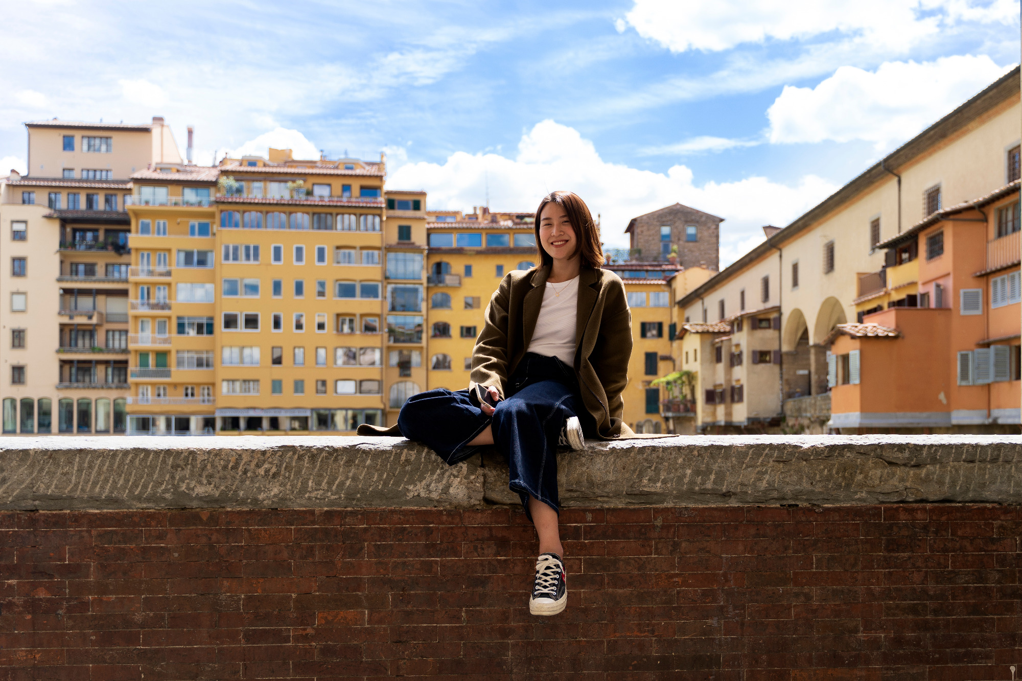 A young woman smiles on a ledge with colorful Florence buildings behind her.