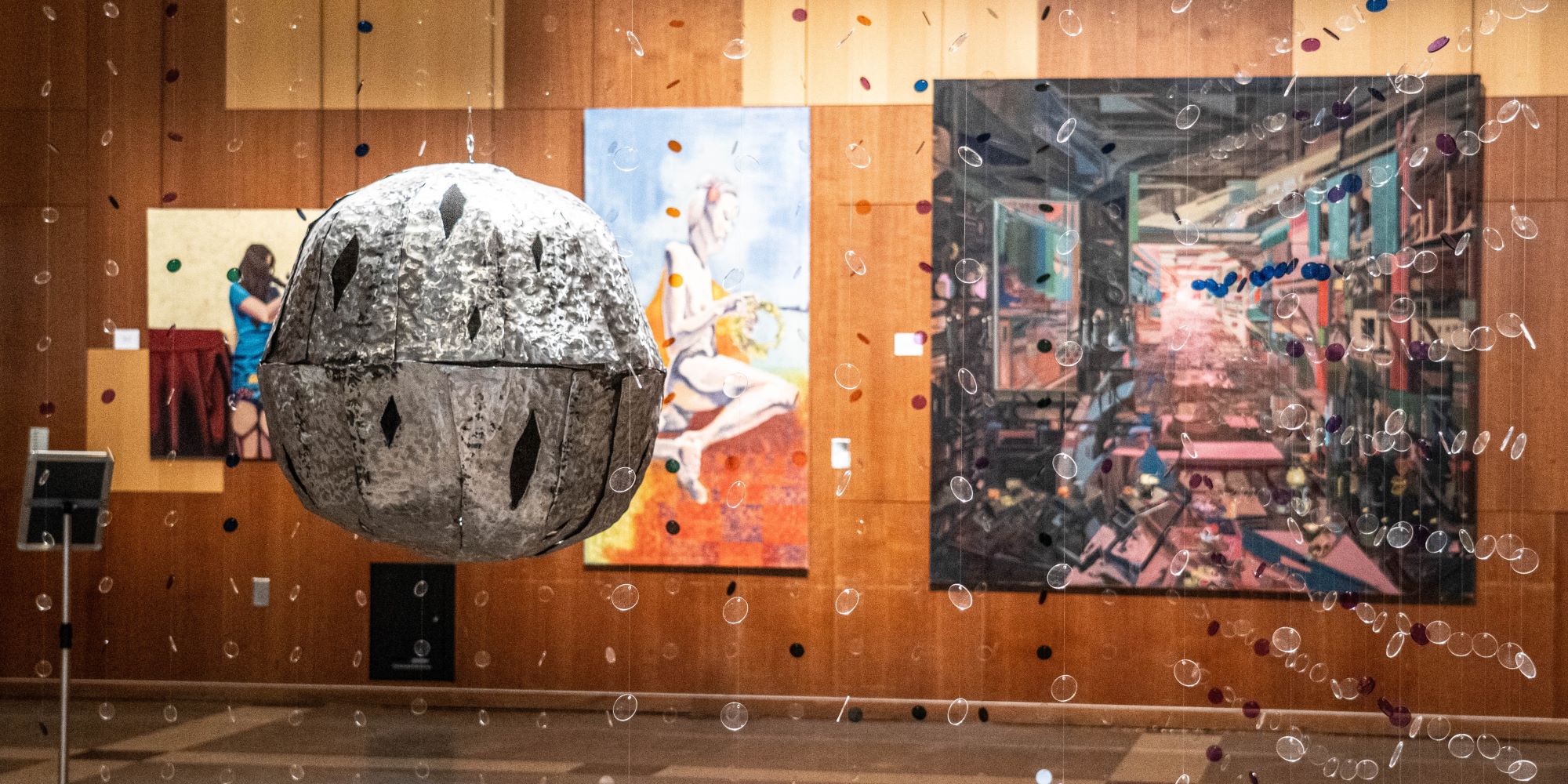 shot of the fine arts student work. Through an installation of multiple strings made of small clear plastic discs in several colors strung on a fishline surrounding an oversized sculpted crotal (jingle) bell, several paintings are visible on the far wall. Discs flash and reflect a projection of small moving lights (reminiscent of a disco ball)
