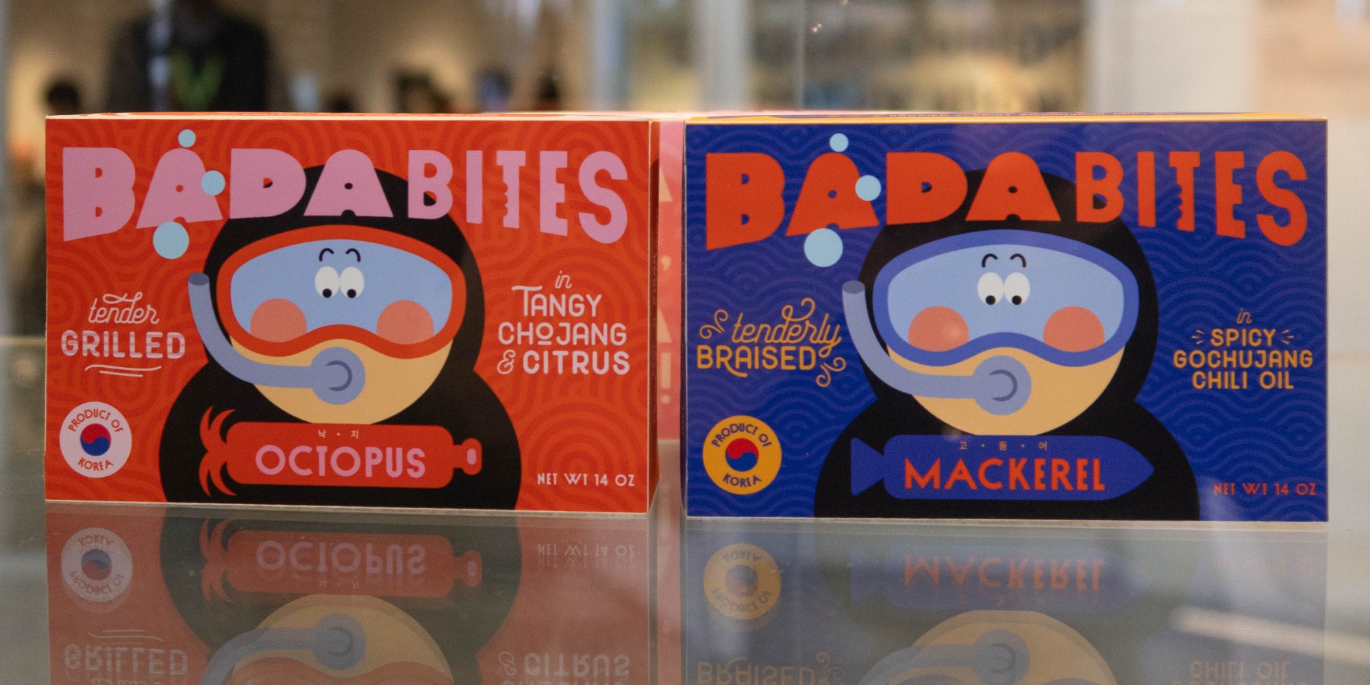 Packaging design vitrine, a close up of two brightly colored boxes for "Bada Bites" seafood products. Blue box with red lettering for mackerel, and red box with pink lettering for octopus. the illustration of a cartoon character with a scuba diving mask and a wet suit prominently appears on both