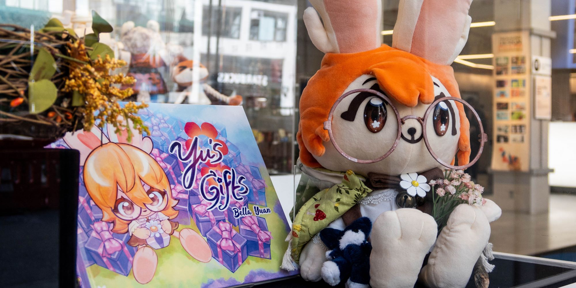 A plush toy of a character with orange hair, bunny ears, and wearing round glasses, which is seated next to an illustrated book, and dried plants. 