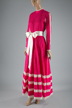 Pink floor-length dress with long sleeves, a wide, white bow at waist and four bands of white lace from hem to knee.