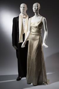 (Left) Men's tuxedo in black wool with matching pants and coordinatind white silk formal scarf with fringe. (Right) floor length evening dress in powder blue silk twill with gold metallic wavy, horizontal sawtooth stripes