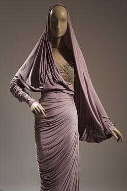 long assymetrical evening sheath inlight purple draped with shirred seams and nude chiffon bodice panel encrusted over left breast with silver and gold beadwork; long right dolman batwing sleeve extending as capelet to left wrist