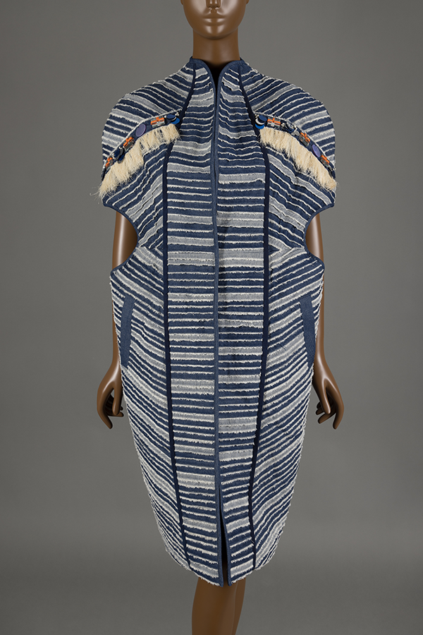 Brown mannequin wearing a blue jacket with striped details of alternating light and dark blue stripes and is decorated with white fringe and black, blue, and orange beads.