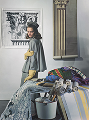 Model Sandra Payson in studio in front of prints of Greek columns, leaning on a table filled with multicolor fabrics wearing a gray cape jacket, yellow gloves, and a grey hat