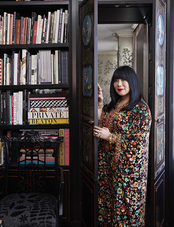 Anna Sui in her home, standing in between two doors next to a floor to ceiling bookshelf filled with books